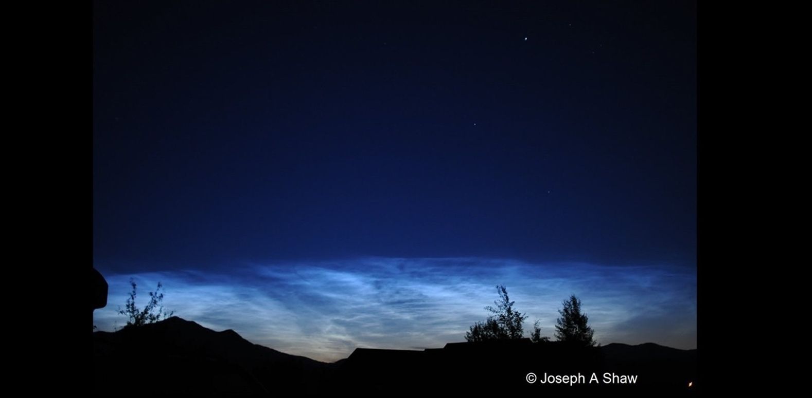 Noctilucent clouds appear to glow against a dark blue sky. The clouds are an electric blue on top, fading to a dusty orange where they dip behind the hills on the horizon.