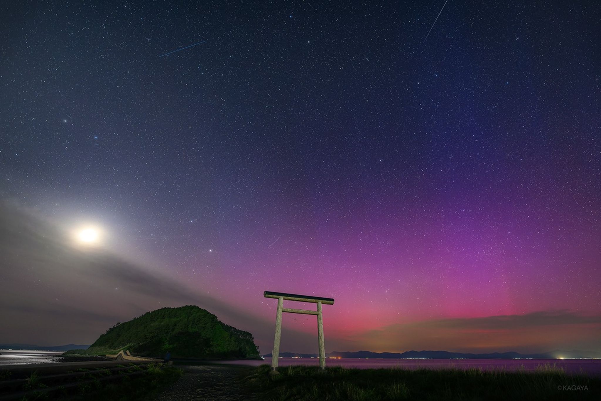 A torii gate stands by the shore with a small hill in the background, under a night sky filled with stars, a bright moon, and vivid purple and pink auroras.